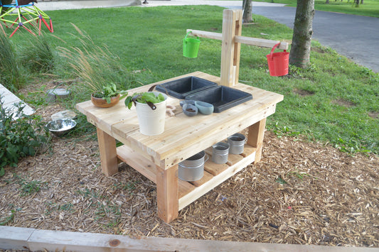 Side view of the cedar Explorer Table with wooden weighing scale, wet-play sink, and removable buckets outdoors.